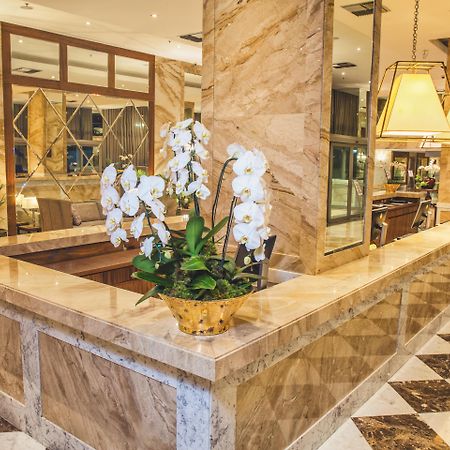 Windsor Marapendi Hotel ริโอเดจาเนโร ภายนอก รูปภาพ The photo depicts a luxurious hotel lobby featuring a polished marble reception desk. There are elegant white orchids arranged in a gold pot on the counter, adding a touch of sophistication. The surrounding interior showcases marble walls, stylish li