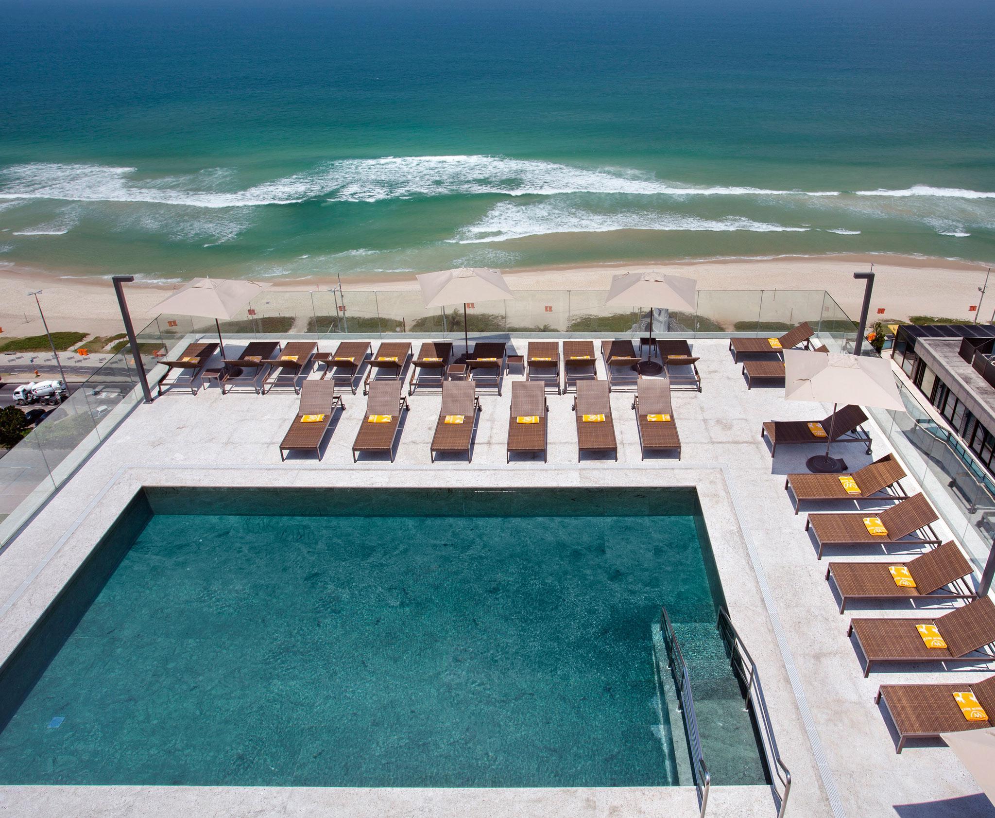 Windsor Marapendi Hotel ริโอเดจาเนโร ภายนอก รูปภาพ The photo shows a rooftop area with a swimming pool and several lounge chairs arranged around it. The pool has a clear blue surface, and the lounge chairs are positioned in a neat row, some with yellow cushions. In the background, you can see a beaut