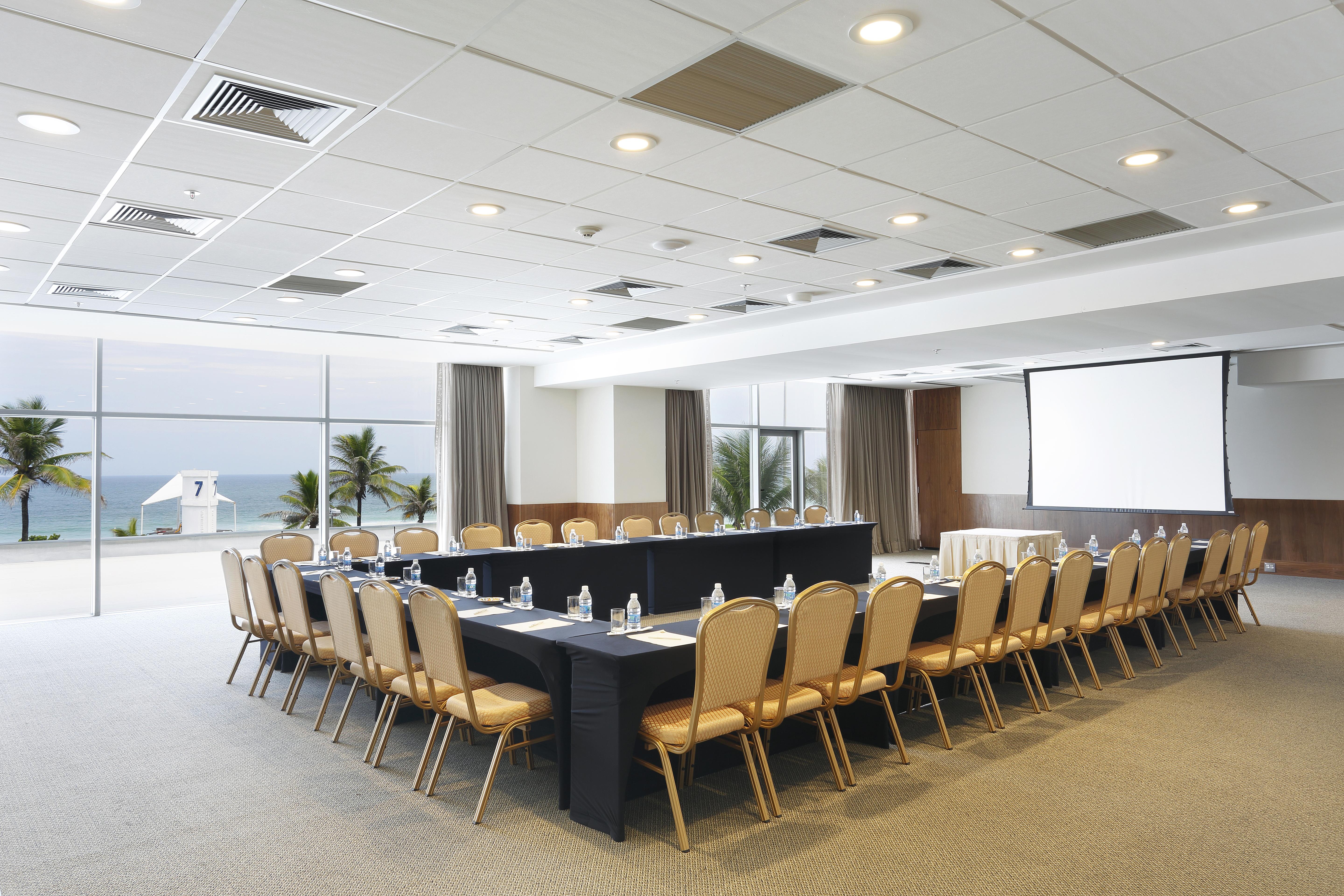 Windsor Marapendi Hotel ริโอเดจาเนโร ภายนอก รูปภาพ The photo shows a spacious conference room set up for a meeting. It features a long, rectangular table surrounded by a number of beige chairs. At the front, there is a large screen, likely for presentations. The room has large windows that provide a 