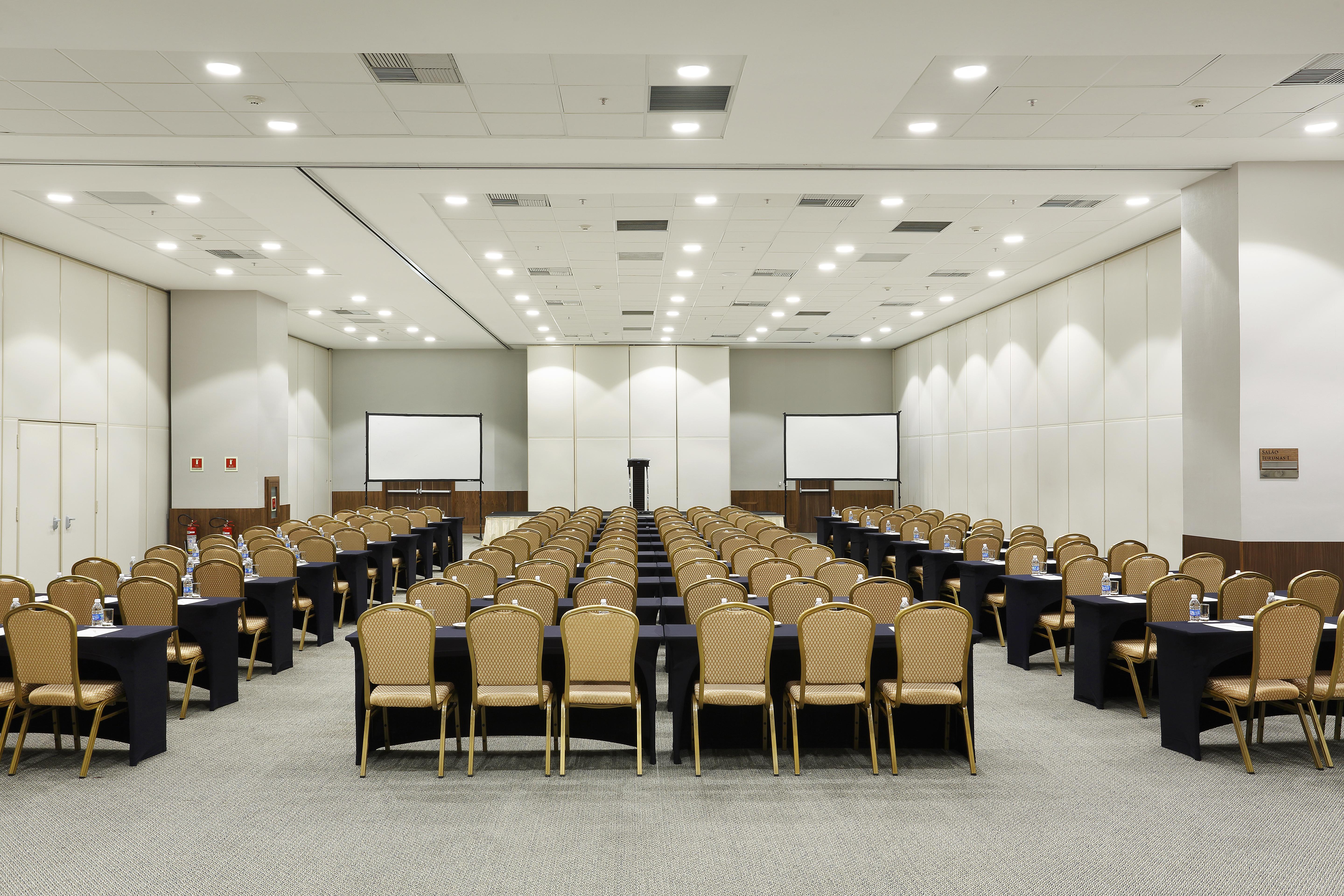 Windsor Marapendi Hotel ริโอเดจาเนโร ภายนอก รูปภาพ The photo shows a conference room arranged for an event or meeting. There are rows of neatly arranged chairs with golden frames, paired with black tables. The room has bright overhead lighting and features two blank screens in the front, likely for p