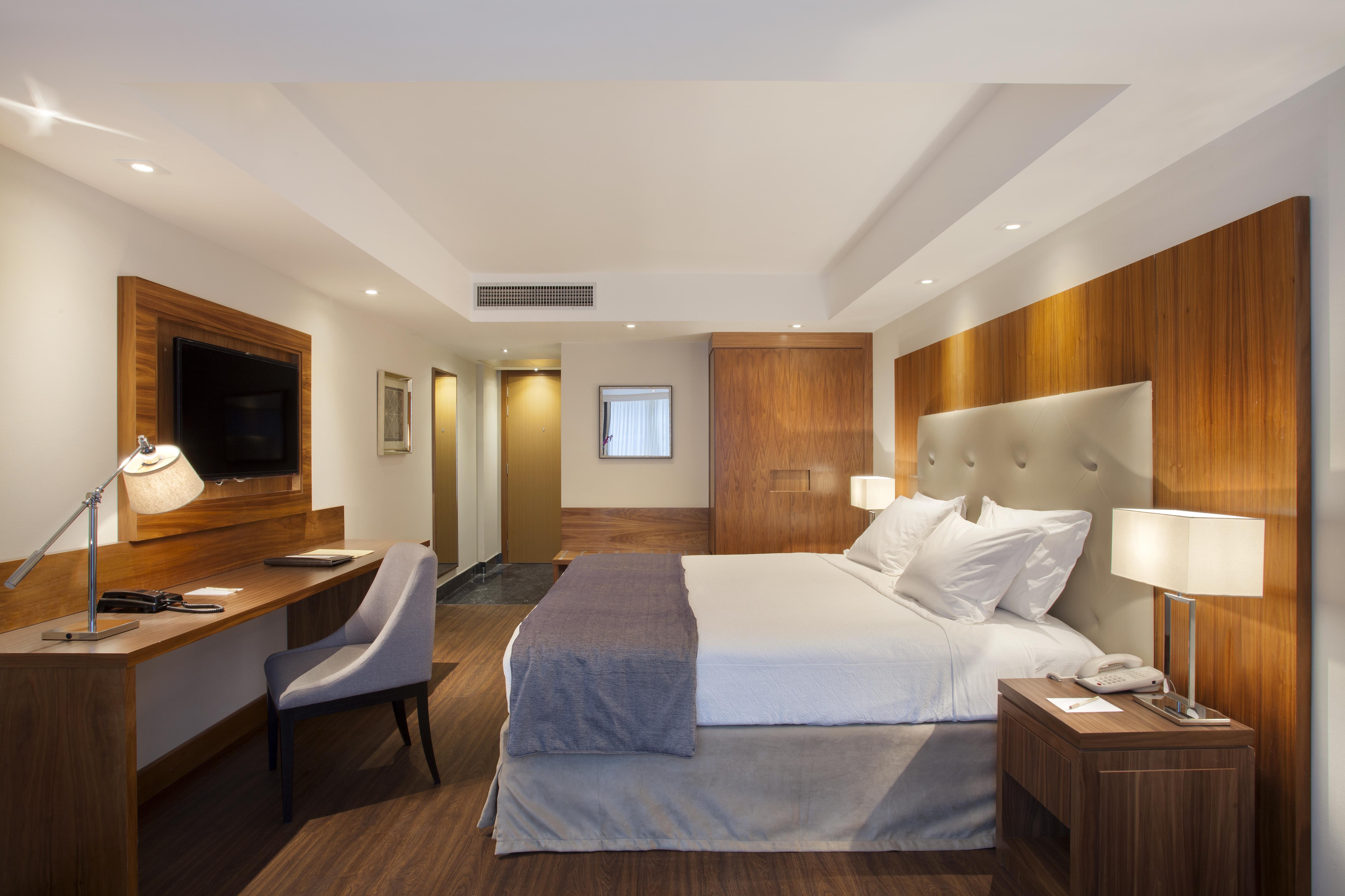 Windsor Marapendi Hotel ริโอเดจาเนโร ภายนอก รูปภาพ The photo shows a modern hotel room featuring a large bed with white and gray bedding. The headboard is upholstered in light fabric. There are two bedside lamps on either side of the bed, providing warm lighting. A television is mounted on the wall o